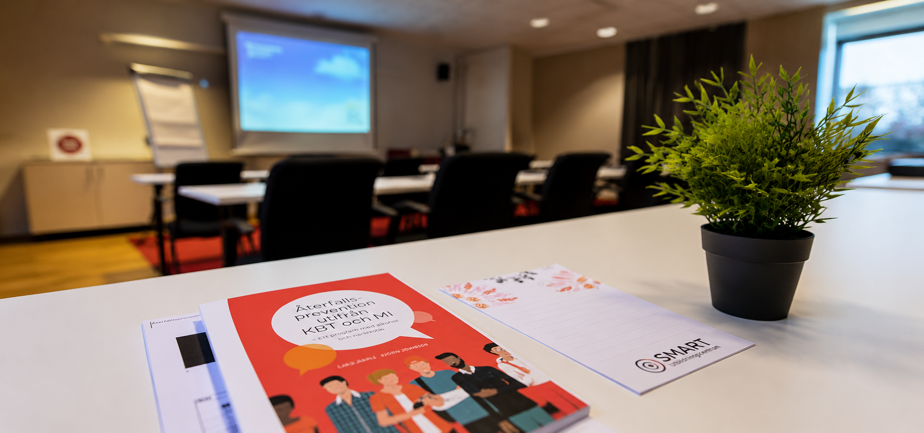 Bok, anteckningsblock och grön växt på bord, med konferensrum och duk projektor i bakgrunden.