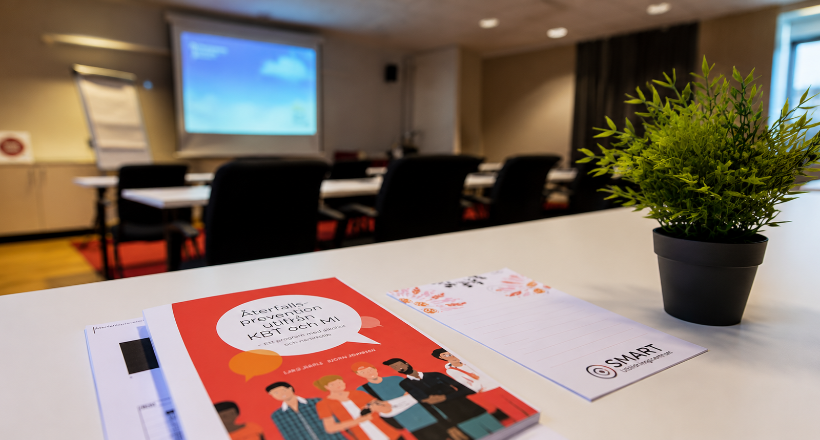 Bok, anteckningsblock och grön växt på bord, med konferensrum och duk projektor i bakgrunden.