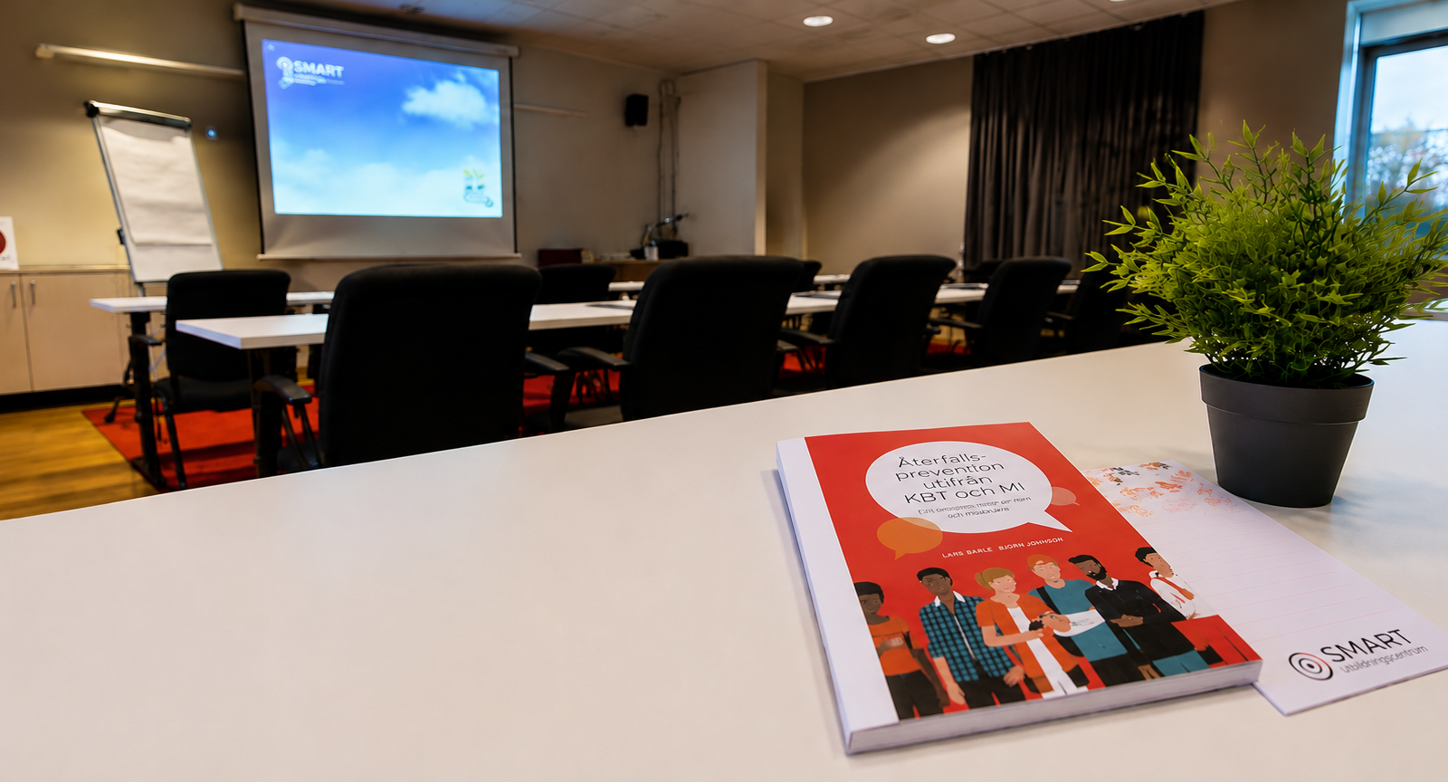 Boken Återfallsprevention utifrån KBT och MI på ett vitt bord i en föreläsningssal med tomma stolar och en projektorduk.