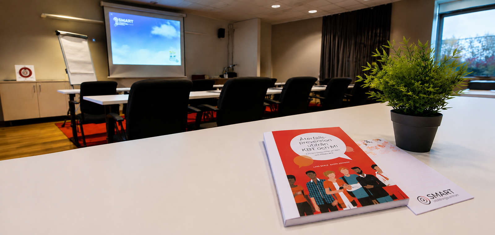 Boken Återfallsprevention utifrån KBT och MI på ett vitt bord i en föreläsningssal med tomma stolar och en projektorduk.