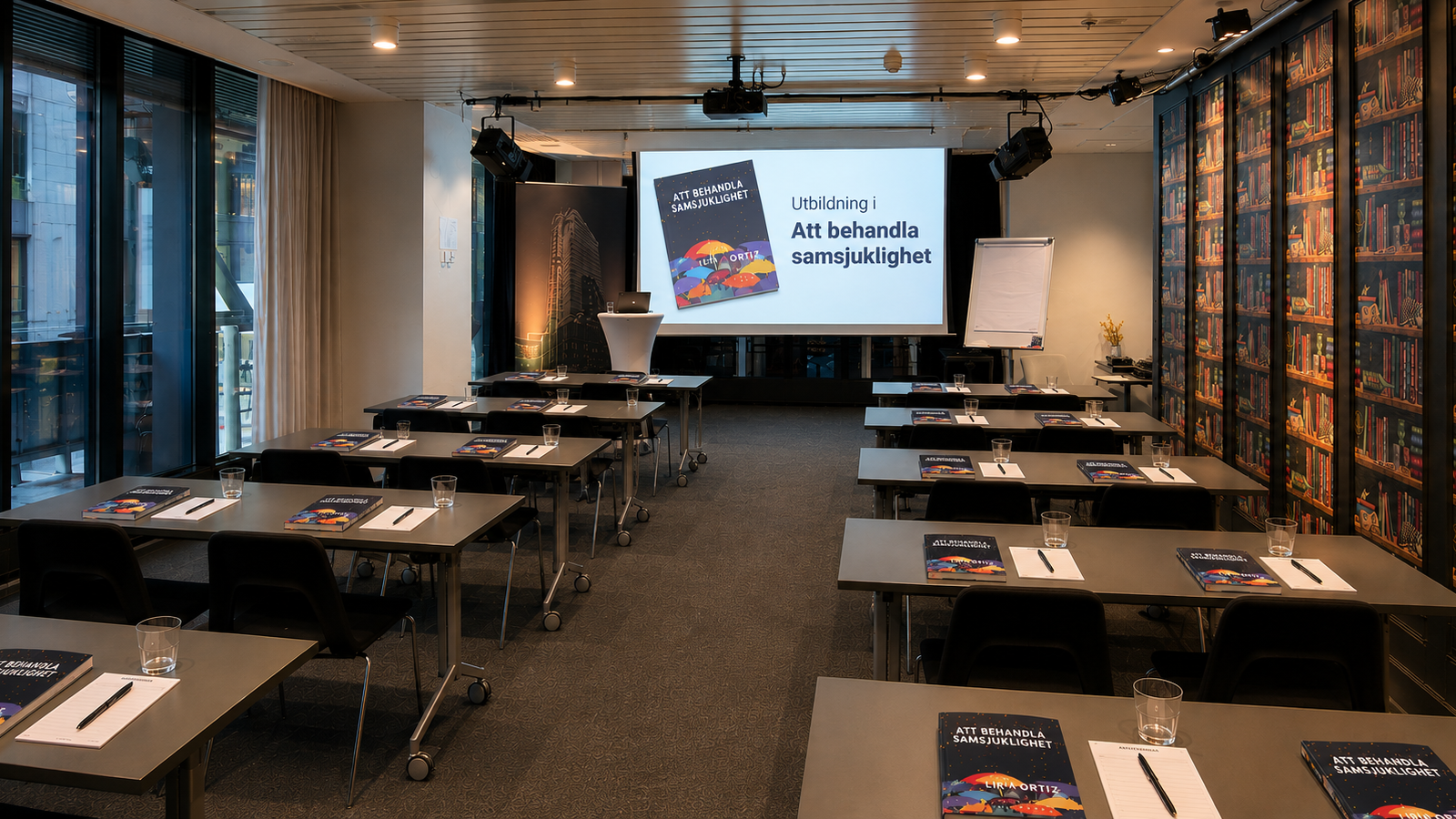 Utbildningslokal med rader av bord, böcker och anteckningsmaterial framför en projektorduk med texten Att behandla samsjuklighet.