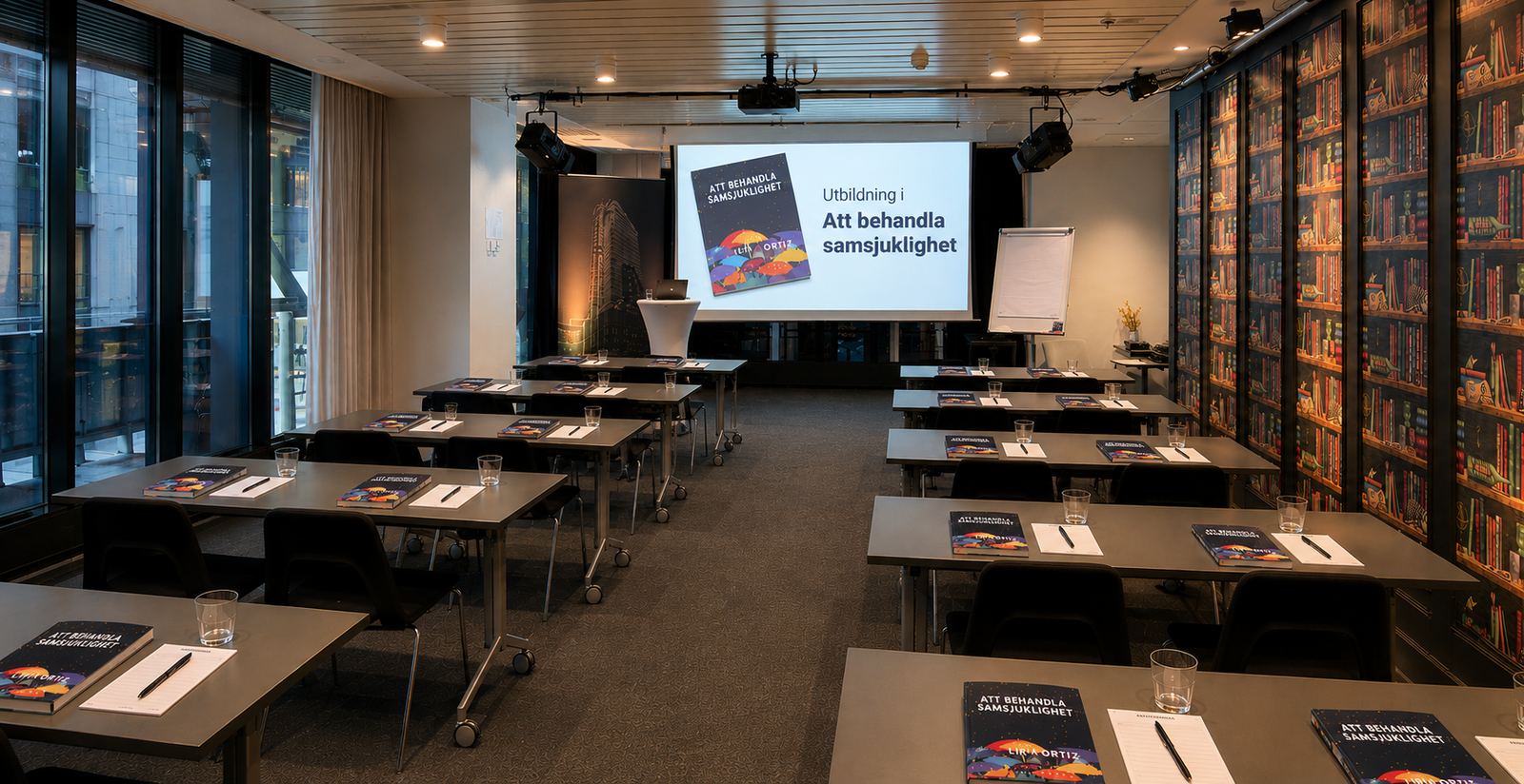 Utbildningslokal med rader av bord, böcker och anteckningsmaterial framför en projektorduk med texten Att behandla samsjuklighet.