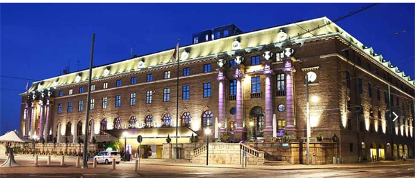 Upplyst fasad på Clarion Hotel Post i tegel med pelare och stentrappa vid entrén under skymningen.
